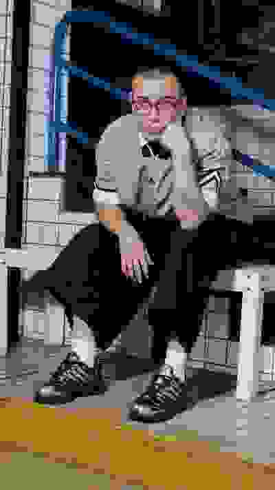 A young person wearing glasses and a uniform sits on a bench in front of a tiled wall.