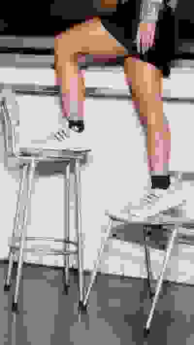 The image shows a person's legs and feet resting on a chair, with the person's lower body visible in the foreground. The background appears to be a tiled floor and a wall.