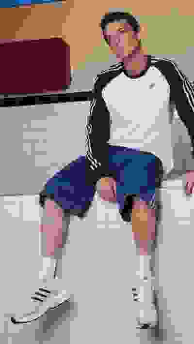 A young man wearing a white and black striped shirt, blue denim shorts, and white sneakers is sitting on a tiled floor in what appears to be a bathroom or kitchen setting.