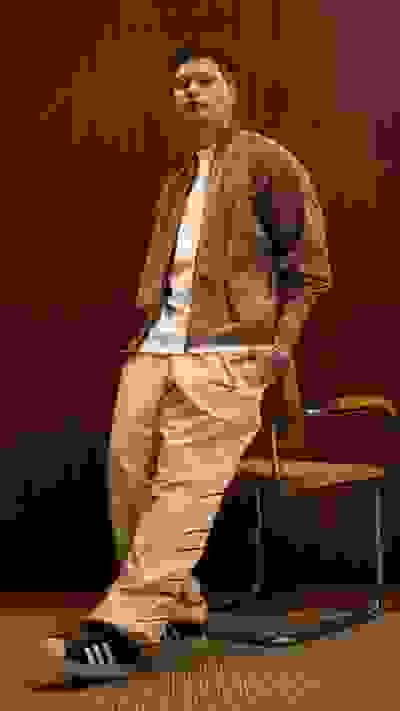 A man in a brown jacket and beige pants is seated on a chair against a wooden background.