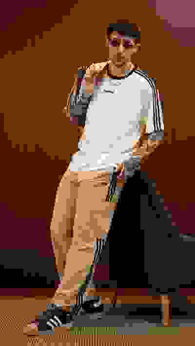 A young person wearing a white shirt, sunglasses, and beige pants is standing in front of a wooden wall.