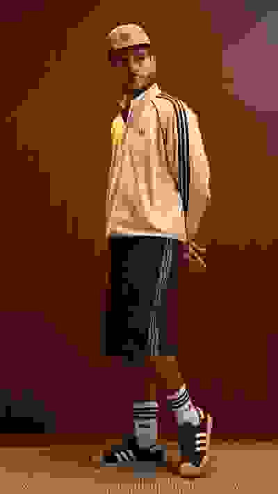 A person wearing a beige jacket, black shorts, and athletic socks stands in front of a wooden wall.