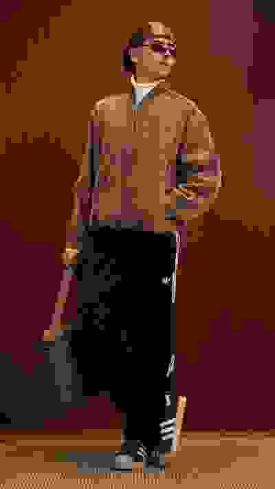 A person wearing a beige jacket, sunglasses, and black pants stands in front of a wooden wall background.