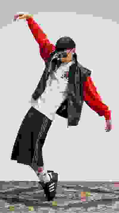 A person in a red jacket and black skirt is jumping and dancing on a patterned rug against a plain background.