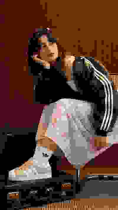 A young woman with curly hair sits on a suitcase, wearing a lace dress and Adidas sneakers, against a wooden background.