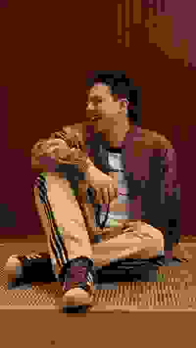 A smiling man in casual clothing sits on the floor, surrounded by a wooden background.