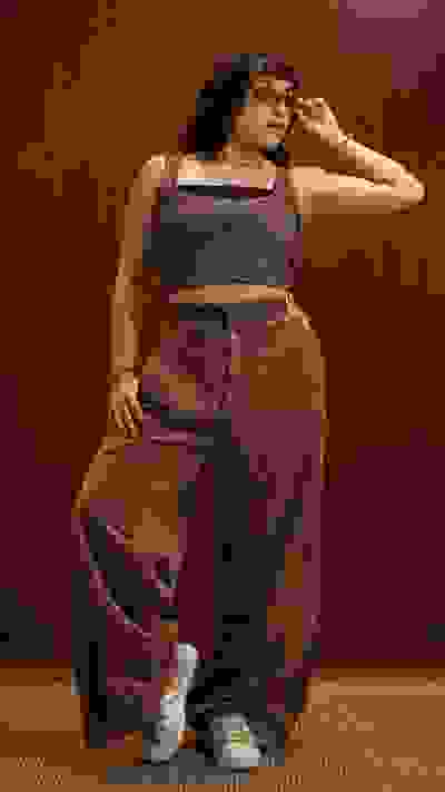 A woman with curly hair wearing a cropped top and wide-leg corduroy pants stands in front of a wooden wall, posing confidently.