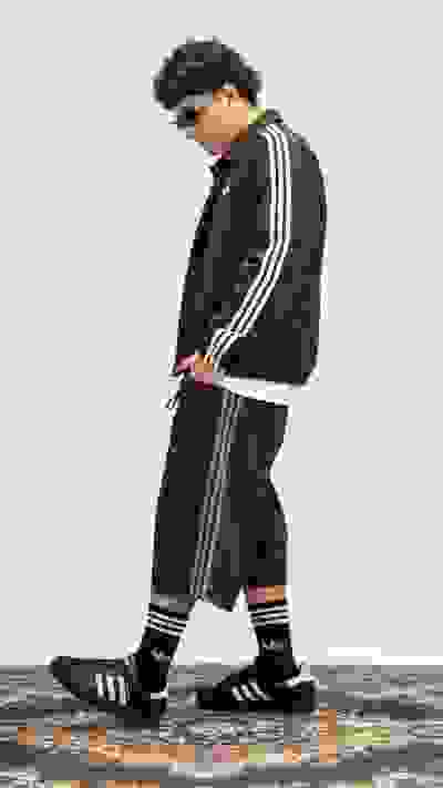 A person wearing a black tracksuit with white stripes stands on a colorful patterned rug against a plain white background.