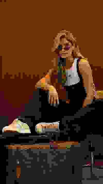 A young woman with long, wavy blonde hair sits on a wooden bench, wearing a black outfit and sunglasses, with a wooden wall in the background.