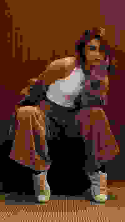 A young woman with long, wavy hair sits on a wooden bench against a plain brown background, wearing a white tank top, beige pants, and white sneakers.