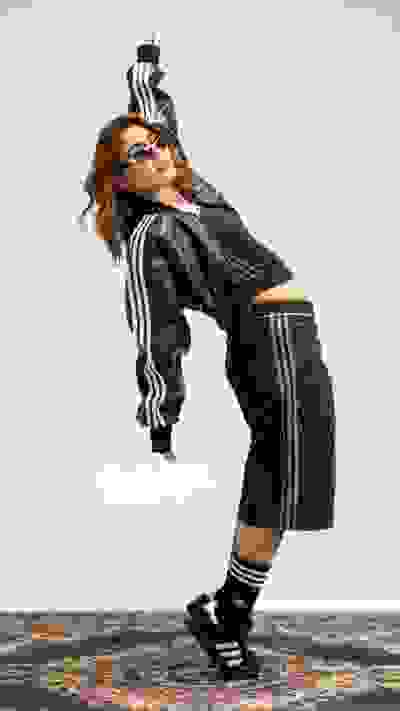 A woman wearing a black and white striped outfit is striking a dynamic pose, with her hair flowing and her body leaning forward, against a plain background.