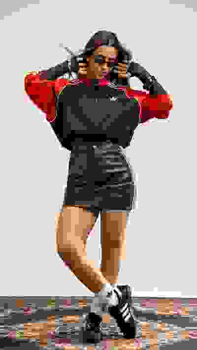 A young woman wearing a red and black tracksuit jacket and a short black skirt stands on a patterned rug, her hair styled in two buns.