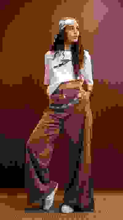 A young woman with curly hair wearing a white top and wide-leg corduroy pants stands in a room with a brown wooden wall.