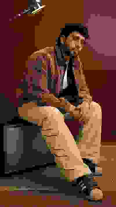 A man with curly hair and a beard sits on a wooden platform, wearing a plaid shirt and beige pants, with a television set in the background.