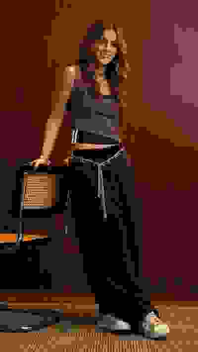 A young woman with wavy hair stands in a room with a wooden wall, holding a guitar case and wearing a patterned top and black pants.