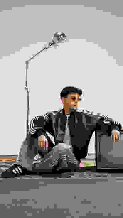 A person wearing a black leather jacket and sunglasses is sitting on a rug, with a lamp and a television set in the background.