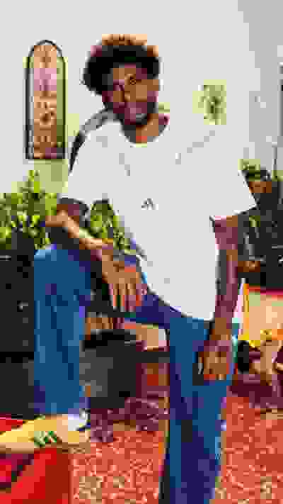 A man with curly hair and tattoos sits on a colorful patterned rug, surrounded by potted plants and a decorative wall hanging in the background.