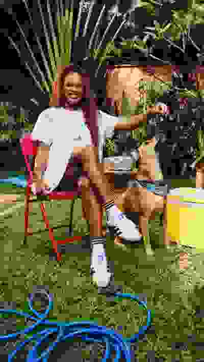 A woman with curly red hair sits in a red chair in a lush, green garden, petting a dog beside her.
