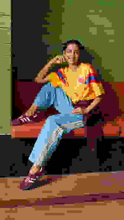 A young woman with a bright, cheerful expression sits on a colorful, patterned couch against a vibrant green wooden wall.