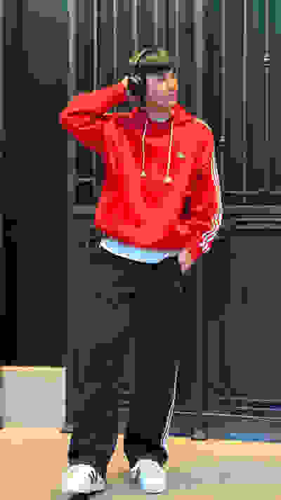 A person wearing a bright red hooded sweatshirt and black pants stands in front of a dark, industrial-looking background with vertical metal panels.