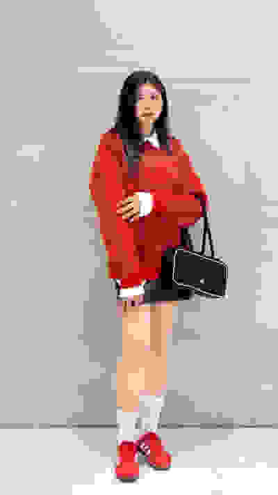 A young woman wearing a bright red sweater, white socks, and red sneakers stands in front of a plain white wall, holding a black handbag.