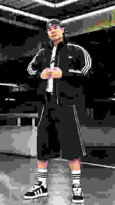 A person wearing a black tracksuit with white stripes, standing in a dimly lit indoor setting with a concrete floor and metal structures in the background.