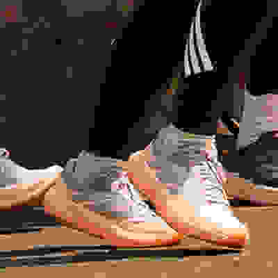The image shows a pair of white and orange sneakers with three stripes on the side, standing on a brick wall background.