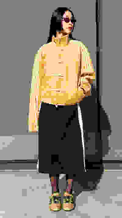 A person wearing a yellow jacket and black skirt stands in front of a gray wall.