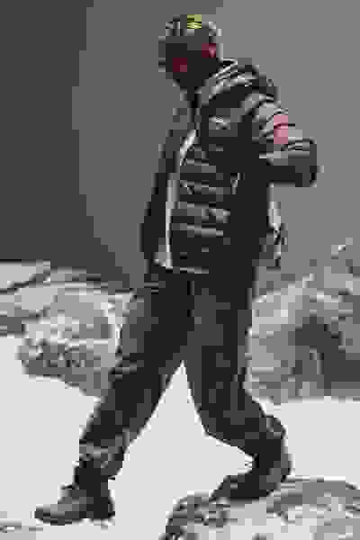 En person, der har en brun dynejakke og grønne bukser på, står på et snedækket, stenet landskab mod en mørk baggrund.