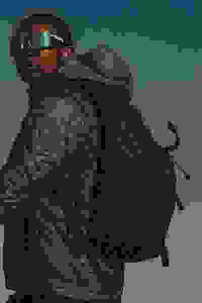 En person, der har en grøn, quiltet jakke og rygsæk med solbriller, står over for en klar blå himmel.