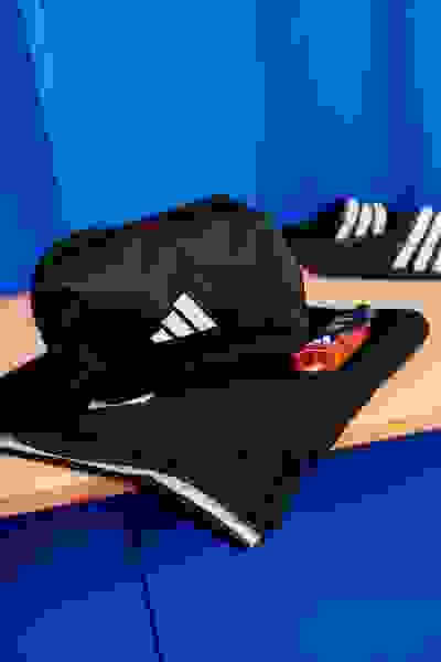 En sort adidas-kasket og et par sorte og hvide adidas-sneakers er placeret på en træoverflade mod en strålende blå baggrund.