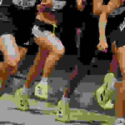 The image shows the lower bodies and legs of several runners wearing bright yellow and green athletic shoes, suggesting a running event or competition taking place in a dimly lit indoor or outdoor setting.