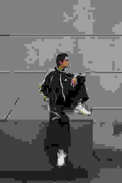 A person wearing a black and green tracksuit is sitting on a concrete structure against a plain gray wall.