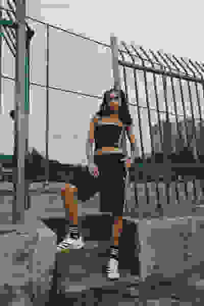 A young woman in a black crop top and shorts is sitting on a concrete structure in front of a metal fence, with buildings visible in the background.