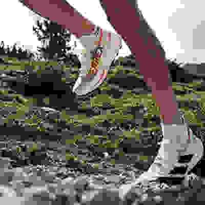 The image shows a person's legs and feet wearing white and orange athletic shoes while running on a rocky, grassy trail in a natural outdoor setting.