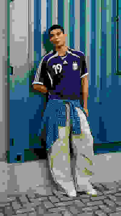 A young man wearing a navy blue sports jersey and khaki pants stands in front of a vibrant blue and white striped wall.