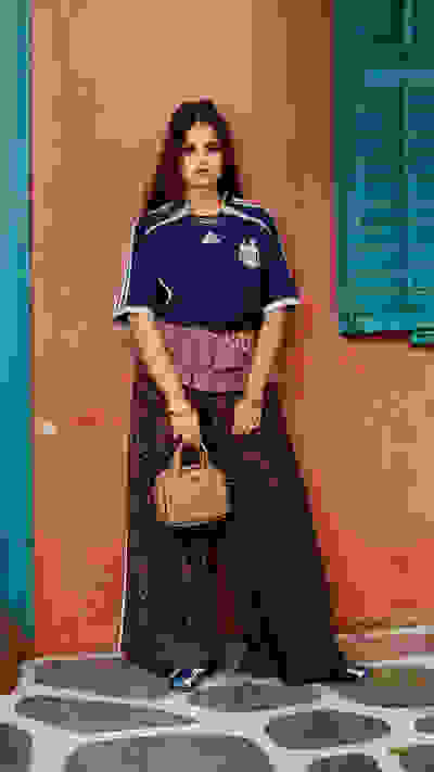 A young woman wearing a blue soccer jersey stands in front of a vibrant pink wall, holding a small handbag and wearing patterned pants and sneakers.