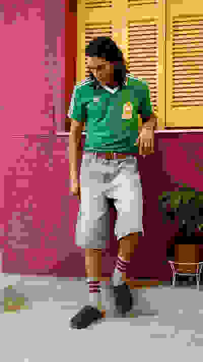 A person wearing a green jersey and light-colored shorts is standing in front of a vibrant pink wall, with potted plants visible in the background.