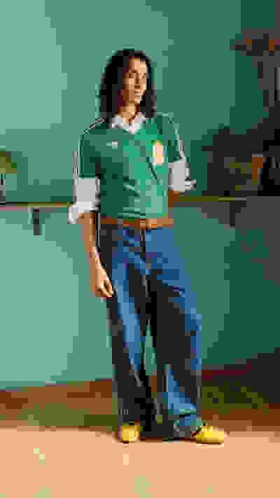 A person wearing a green jersey, blue jeans, and yellow shoes stands in a room with a green wall and shelves in the background.