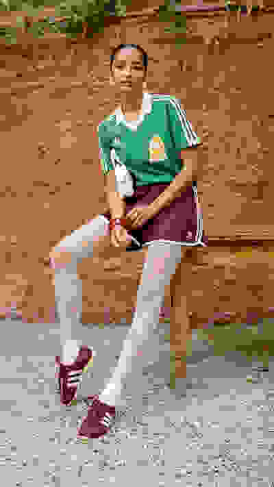 A young person wearing a green sports jersey and burgundy shorts is sitting on a wooden bench in front of a brick wall with vines growing on it.