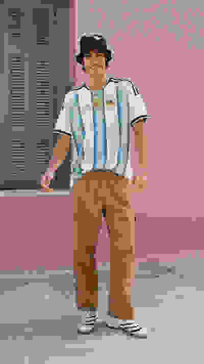 A young man wearing an Argentina national team jersey, a black hat, and beige pants stands in front of a pink-colored building with louvered windows.