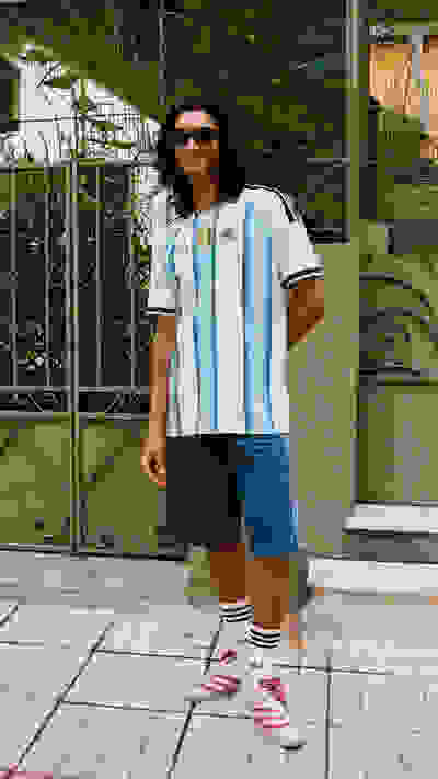 The image shows a person wearing a light blue and white jersey, blue shorts, and white sneakers, standing in front of a decorative metal gate with a green background.
