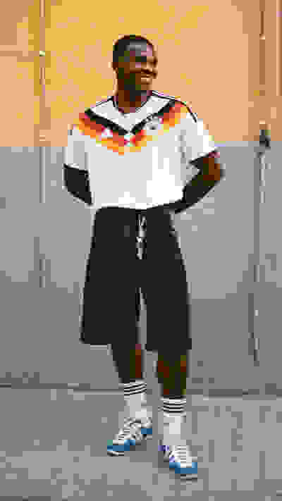 A man wearing a white and colorful jersey stands in front of a yellow wall, with his hands in his pockets and a confident expression on his face.