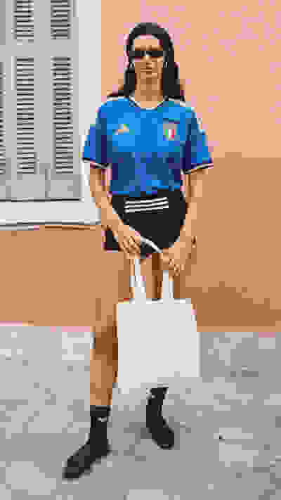 A person wearing a blue sports jersey and holding a white tote bag stands in front of a pink-colored building with shuttered windows.