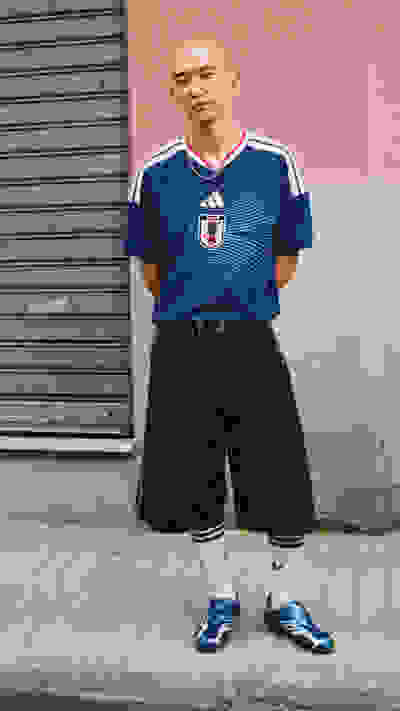 A person wearing a blue and white sports jersey, black shorts, and blue and white sneakers stands in front of a pink wall with a metal shutter in the background.