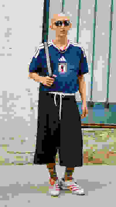 A person wearing a blue and white sports jersey, black shorts, and red and white sneakers stands in front of a weathered, blue-painted wall.