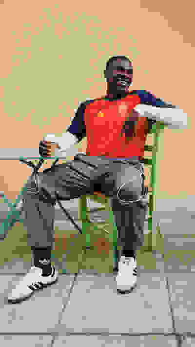 A man wearing an orange jersey and casual clothing is sitting on a colorful chair, holding a cup and appearing relaxed in an outdoor setting with a plain background.