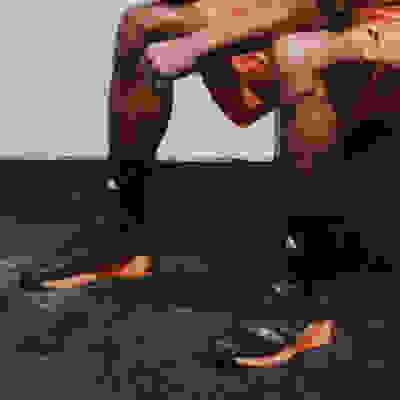 The image shows a person wearing orange athletic clothing and black and orange sneakers, sitting on a dark surface with a body of water in the background.