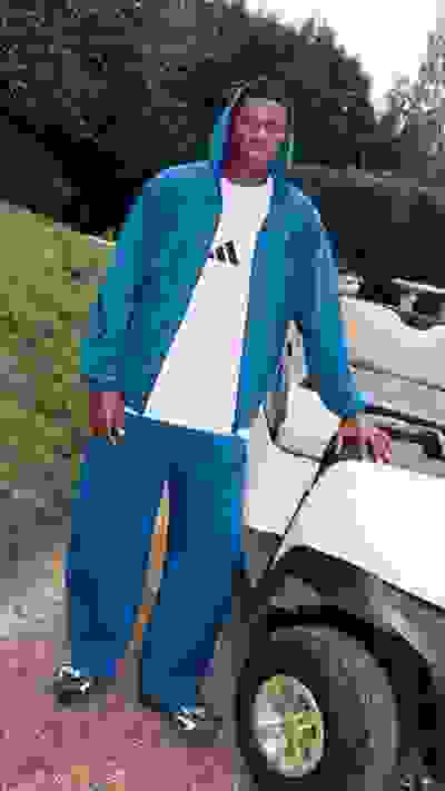 A person wearing a blue hooded jacket and pants is standing in front of a vehicle on a grassy area surrounded by hedges.