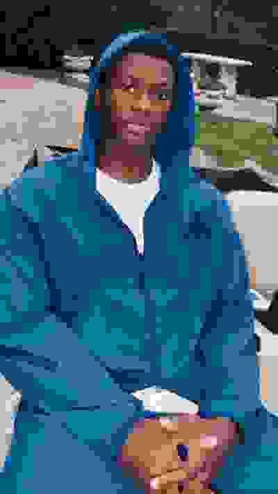 A young man wearing a blue hooded jacket is sitting on a bench in an outdoor setting with greenery and a stone structure visible in the background.